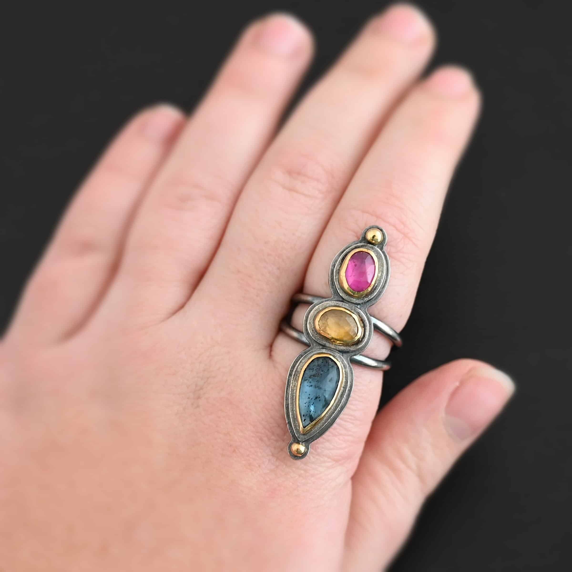 Big & Awesome Ring Close up of the "Big and Awesome" Ring. Made out of sterling silver, 18k gold, ruby, yellow sapphire, and kyanite. Ring has two separate bands that join together at the back. The kyanite is the top stone, and the largest. It is a teardrop shape with the point facing upwards. It is a blue color with black speckles throughout the ring. The yellow sapphire is the middle stone. It is an oval shape. It is a dark yellow color. The final stone is a ruby. It is an oval shape as well, but it is set vertically instead of horizontally. It is a dark pink color. There are gold dots on the bottom and top of the ring. Each stone is set in gold. Ring is worn on the middle finger of a models hand to demonstrate how long it is.