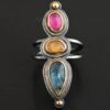 Close up of the "Big and Awesome" Ring. Made out of sterling silver, 18k gold, ruby, yellow sapphire, and kyanite. Ring has two separate bands that join together at the back. The kyanite is the top stone, and the largest. It is a teardrop shape with the point facing upwards. It is a blue color with black speckles throughout the ring. The yellow sapphire is the middle stone. It is an oval shape. It is a dark yellow color. The final stone is a ruby. It is an oval shape as well, but it is set vertically instead of horizontally. It is a dark pink color. There are gold dots on the bottom and top of the ring. Each stone is set in gold. Ring is set on a leather stand so it stays upward. The leather stand is set against a dark background.