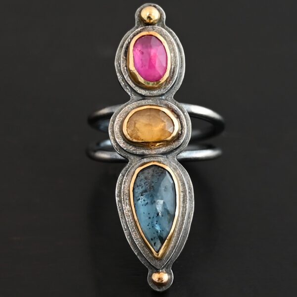 Close up of the "Big and Awesome" Ring. Made out of sterling silver, 18k gold, ruby, yellow sapphire, and kyanite. Ring has two separate bands that join together at the back. The kyanite is the top stone, and the largest. It is a teardrop shape with the point facing upwards. It is a blue color with black speckles throughout the ring. The yellow sapphire is the middle stone. It is an oval shape. It is a dark yellow color. The final stone is a ruby. It is an oval shape as well, but it is set vertically instead of horizontally. It is a dark pink color. There are gold dots on the bottom and top of the ring. Each stone is set in gold. Ring is set on a leather stand so it stays upward. The leather stand is set against a dark background.