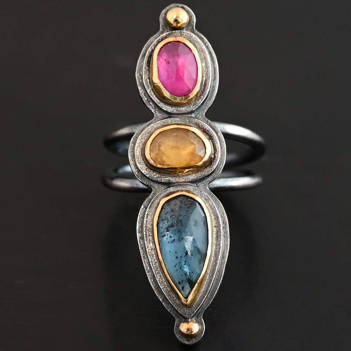 Big & Awesome Ring New Close up of the "Big and Awesome" Ring. Made out of sterling silver, 18k gold, ruby, yellow sapphire, and kyanite. Ring has two separate bands that join together at the back. The kyanite is the top stone, and the largest. It is a teardrop shape with the point facing upwards. It is a blue color with black speckles throughout the ring. The yellow sapphire is the middle stone. It is an oval shape. It is a dark yellow color. The final stone is a ruby. It is an oval shape as well, but it is set vertically instead of horizontally. It is a dark pink color. There are gold dots on the bottom and top of the ring. Each stone is set in gold. Ring is set on a leather stand so it stays upward. The leather stand is set against a dark background.