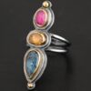 Close up of the "Big and Awesome" Ring. Made out of sterling silver, 18k gold, ruby, yellow sapphire, and kyanite. Ring has two separate bands that join together at the back. The kyanite is the top stone, and the largest. It is a teardrop shape with the point facing upwards. It is a blue color with black speckles throughout the ring. The yellow sapphire is the middle stone. It is an oval shape. It is a dark yellow color. The final stone is a ruby. It is an oval shape as well, but it is set vertically instead of horizontally. It is a dark pink color. There are gold dots on the bottom and top of the ring. Each stone is set in gold. Ring is set on a leather stand so it stays upward. The leather stand is set against a dark background.