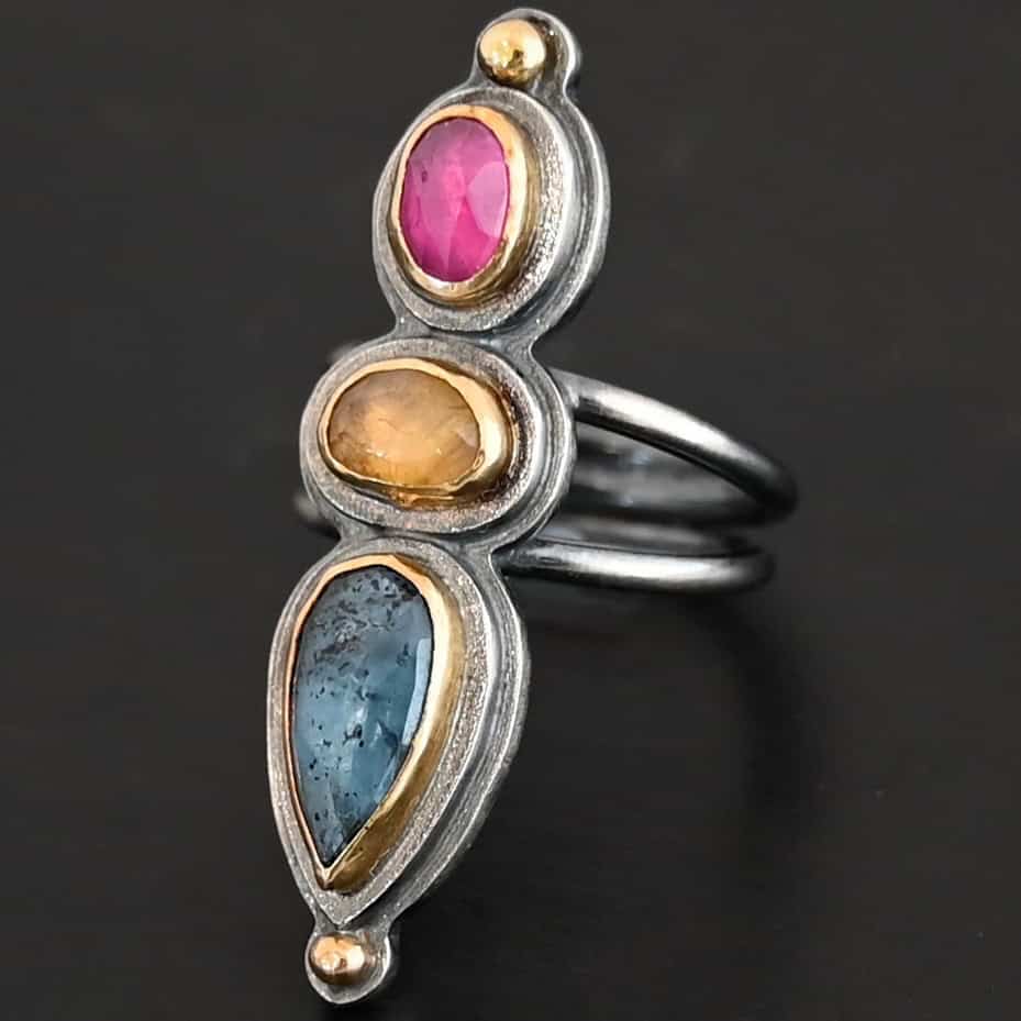 Big & Awesome Ring New Close up of the "Big and Awesome" Ring. Made out of sterling silver, 18k gold, ruby, yellow sapphire, and kyanite. Ring has two separate bands that join together at the back. The kyanite is the top stone, and the largest. It is a teardrop shape with the point facing upwards. It is a blue color with black speckles throughout the ring. The yellow sapphire is the middle stone. It is an oval shape. It is a dark yellow color. The final stone is a ruby. It is an oval shape as well, but it is set vertically instead of horizontally. It is a dark pink color. There are gold dots on the bottom and top of the ring. Each stone is set in gold. Ring is set on a leather stand so it stays upward. The leather stand is set against a dark background.