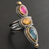 Close up of the "Big and Awesome" Ring. Made out of sterling silver, 18k gold, ruby, yellow sapphire, and kyanite. Ring has two separate bands that join together at the back. The kyanite is the top stone, and the largest. It is a teardrop shape with the point facing upwards. It is a blue color with black speckles throughout the ring. The yellow sapphire is the middle stone. It is an oval shape. It is a dark yellow color. The final stone is a ruby. It is an oval shape as well, but it is set vertically instead of horizontally. It is a dark pink color. There are gold dots on the bottom and top of the ring. Each stone is set in gold. Ring is set on a leather stand so it stays upward. The leather stand is set against a dark background.