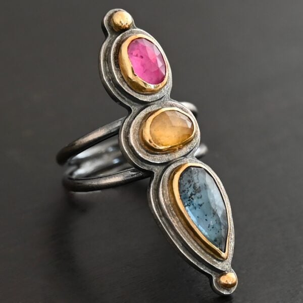 Close up of the "Big and Awesome" Ring. Made out of sterling silver, 18k gold, ruby, yellow sapphire, and kyanite. Ring has two separate bands that join together at the back. The kyanite is the top stone, and the largest. It is a teardrop shape with the point facing upwards. It is a blue color with black speckles throughout the ring. The yellow sapphire is the middle stone. It is an oval shape. It is a dark yellow color. The final stone is a ruby. It is an oval shape as well, but it is set vertically instead of horizontally. It is a dark pink color. There are gold dots on the bottom and top of the ring. Each stone is set in gold. Ring is set on a leather stand so it stays upward. The leather stand is set against a dark background.