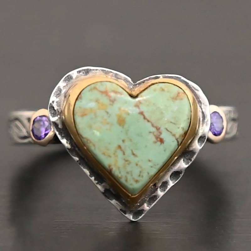 Close up of the "Wild Beauty" Ring. Made out of sterling silver, gold, turquoise, and amethyst. The main stone is a heart shaped turquoise that is set in gold. The turquoise is a mint green color, with lines of gold and brown flowing throughout it. There is a small circular amethyst that is set in gold on the left side of the ring. The sterling silver band is engraved with leaves. Ring is set against a dark background.