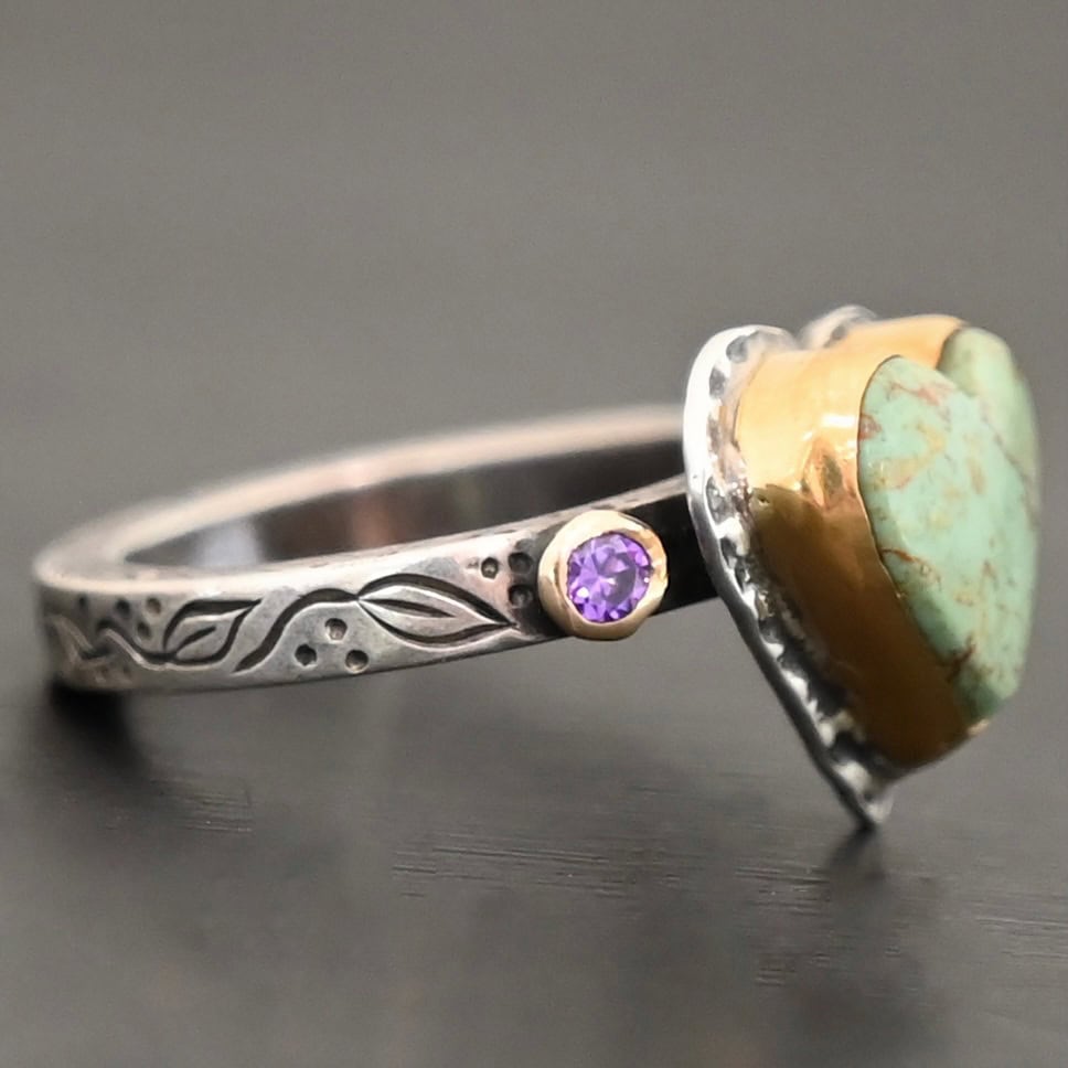 Close up of the "Wild Beauty" Ring. Made out of sterling silver, gold, turquoise, and amethyst. The main stone is a heart shaped turquoise that is set in gold. The turquoise is a mint green color, with lines of gold and brown flowing throughout it. There is a small circular amethyst that is set in gold on the left side of the ring. The sterling silver band is engraved with leaves. Ring is set against a dark background.