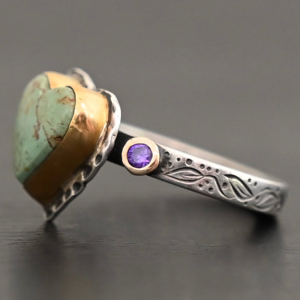 Close up of the "Wild Beauty" Ring. Made out of sterling silver, gold, turquoise, and amethyst. The main stone is a heart shaped turquoise that is set in gold. The turquoise is a mint green color, with lines of gold and brown flowing throughout it. There is a small circular amethyst that is set in gold on the left side of the ring. The sterling silver band is engraved with leaves. Ring is set against a dark background.