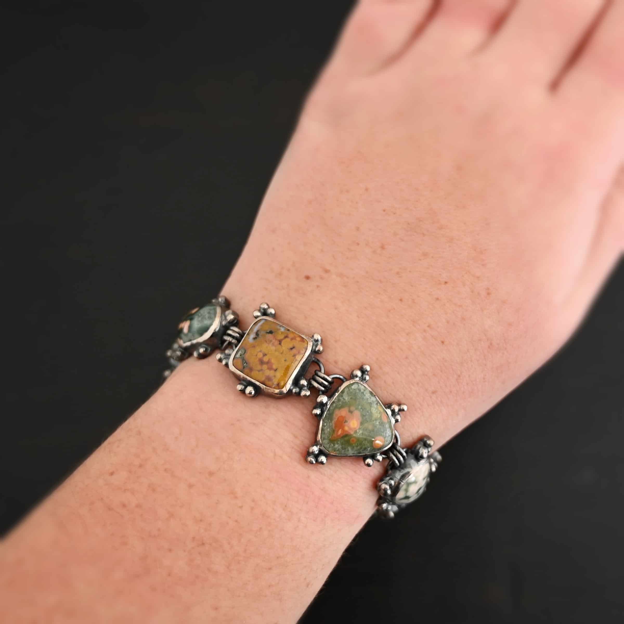 Life Is A Painting Bracelet Close up of the "Life Is A Painting" Bracelet. Made out of sterling silver and ocean jasper. The ocean jasper are a variety of colors, but primarily dark yellow, light and dark green, and red. They are a variety of shapes. The bracelet comes with a toggle clasp. Each stone is connected closely with three jump rings, and each stone setting has sterling silver dots scattered around it. Bracelet is set on the fingers of a hand to demonstrate size of the stones. Bracelet is worn by a model to demonstrate size of the stones.