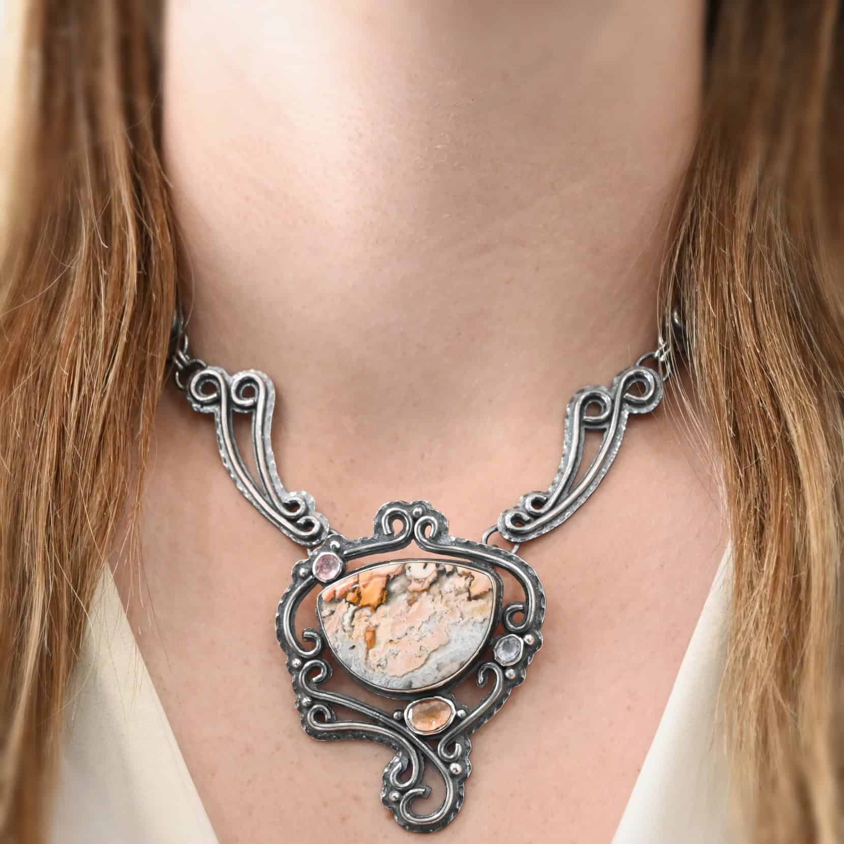 Meadow Wind Necklace The "Meadow Wind" Necklace as shown on a model. Necklace is made out of sterling silver, crazy lace agate, sapphires, and tourmaline. The agate is the main stone, and it is a slight oval shape. There are three other stones that are scattered around the necklace. The agate is surrounded by sterling silver swirls, and the collar of the necklace is made out of these swirls as well. Necklace is worn by a model wearing a white v-neck shirt.
