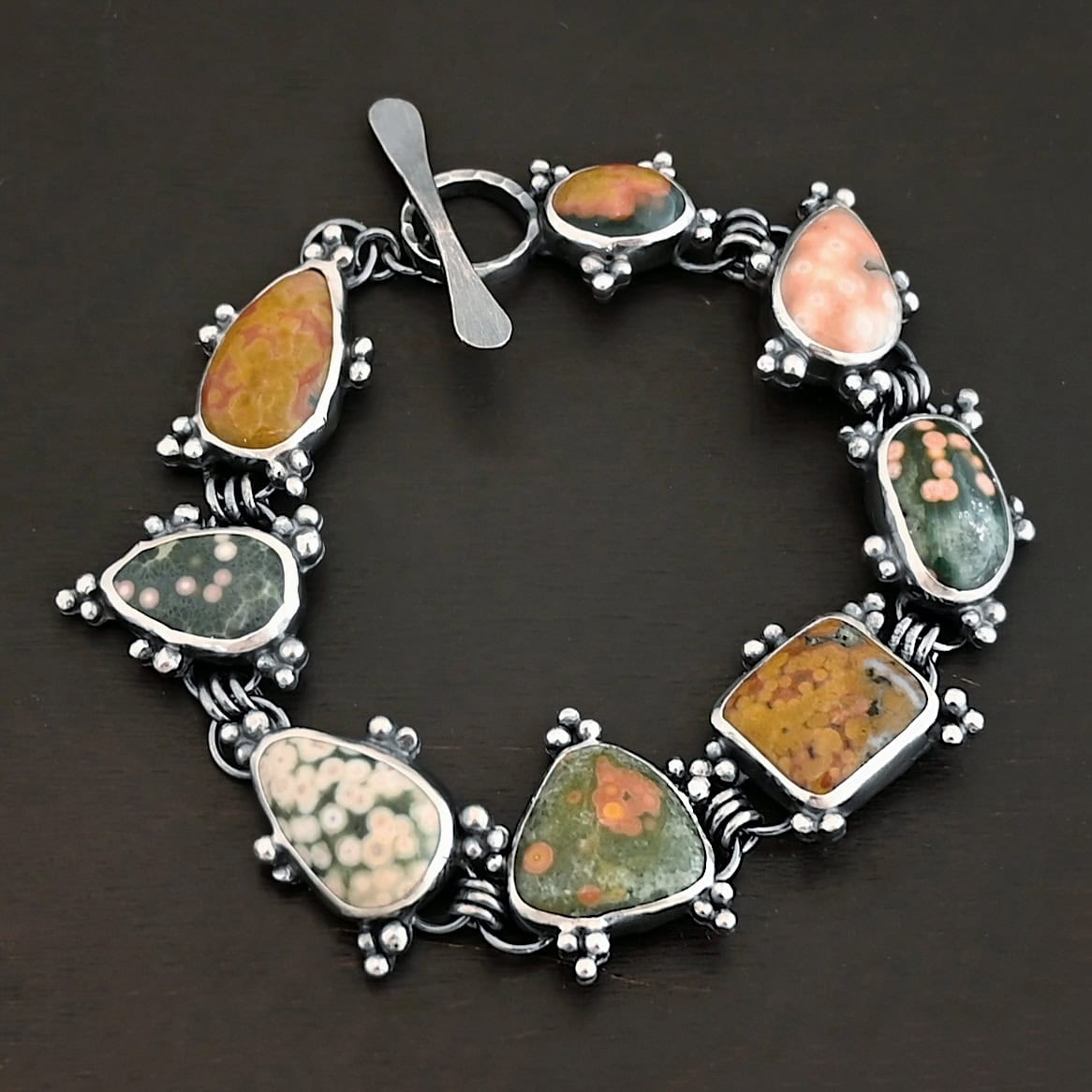 Life Is A Painting Bracelet Close up of the "Life Is A Painting" Bracelet. Made out of sterling silver and ocean jasper. The ocean jasper are a variety of colors, but primarily dark yellow, light and dark green, and light pink. They are a variety of shapes. The bracelet comes with a toggle clasp. Each stone is connected closely with three jump rings, and each stone setting has sterling silver dots scattered around it. Bracelet is set against a dark background.