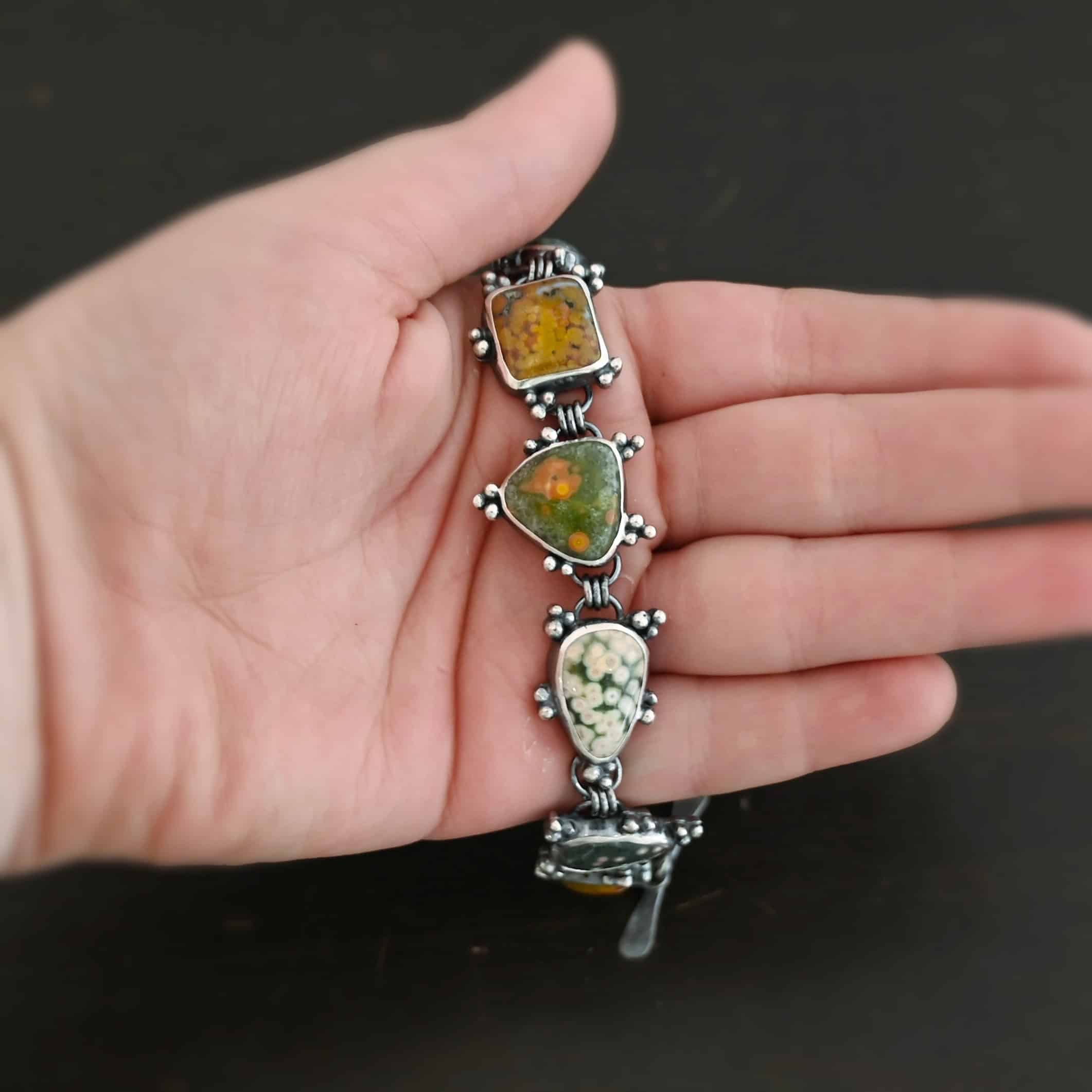 Life Is A Painting Bracelet Close up of the "Life Is A Painting" Bracelet. Made out of sterling silver and ocean jasper. The ocean jasper are a variety of colors, but primarily dark yellow, light and dark green, and red. They are a variety of shapes. The bracelet comes with a toggle clasp. Each stone is connected closely with three jump rings, and each stone setting has sterling silver dots scattered around it. Bracelet is set on the fingers of a hand to demonstrate size of the stones. Bracelet is in the palm of a hand to demonstrate size of the stones.