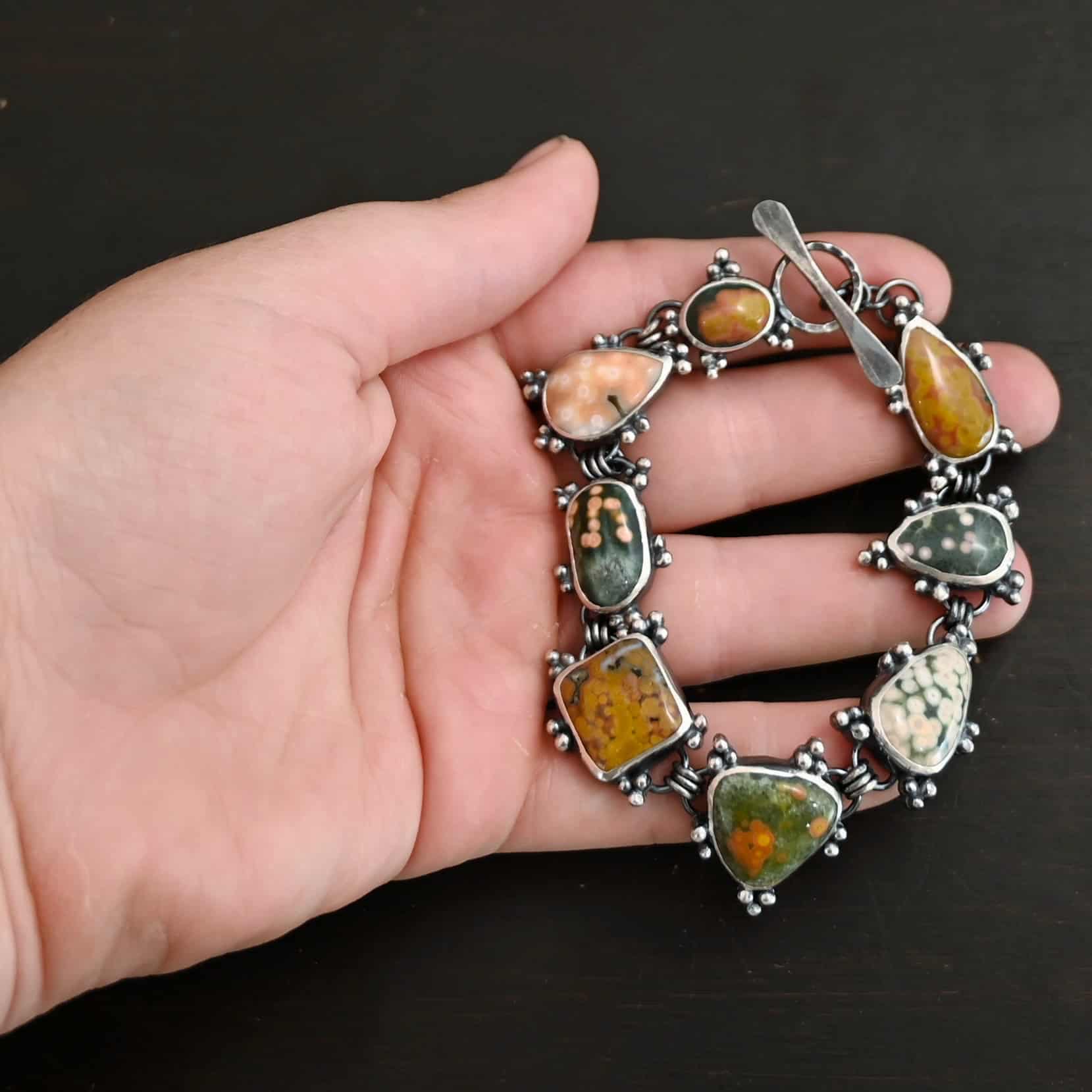 Life Is A Painting Bracelet Close up of the "Life Is A Painting" Bracelet. Made out of sterling silver and ocean jasper. The ocean jasper are a variety of colors, but primarily dark yellow, light and dark green, and light pink. They are a variety of shapes. The bracelet comes with a toggle clasp. Each stone is connected closely with three jump rings, and each stone setting has sterling silver dots scattered around it. Bracelet is set on the fingers of a hand to demonstrate size of the stones. Hand is resting against a dark background.