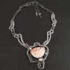 The "Meadow Wind" Necklace as shown close up. Necklace is made out of sterling silver, crazy lace agate, sapphires, and tourmaline. The agate is the main stone, and it is a slight oval shape. There are three other stones that are scattered around the necklace. The agate is surrounded by sterling silver swirls, and the collar of the necklace is made out of these swirls as well. Necklace is set against a dark background.
