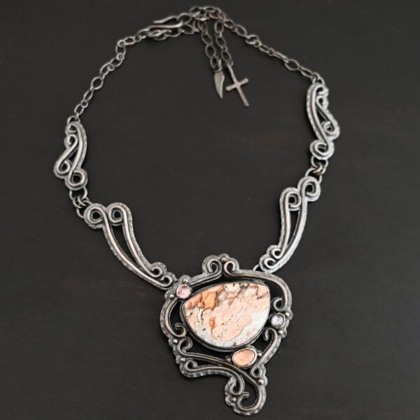 The "Meadow Wind" Necklace as shown close up. Necklace is made out of sterling silver, crazy lace agate, sapphires, and tourmaline. The agate is the main stone, and it is a slight oval shape. There are three other stones that are scattered around the necklace. The agate is surrounded by sterling silver swirls, and the collar of the necklace is made out of these swirls as well. Necklace is set against a dark background.