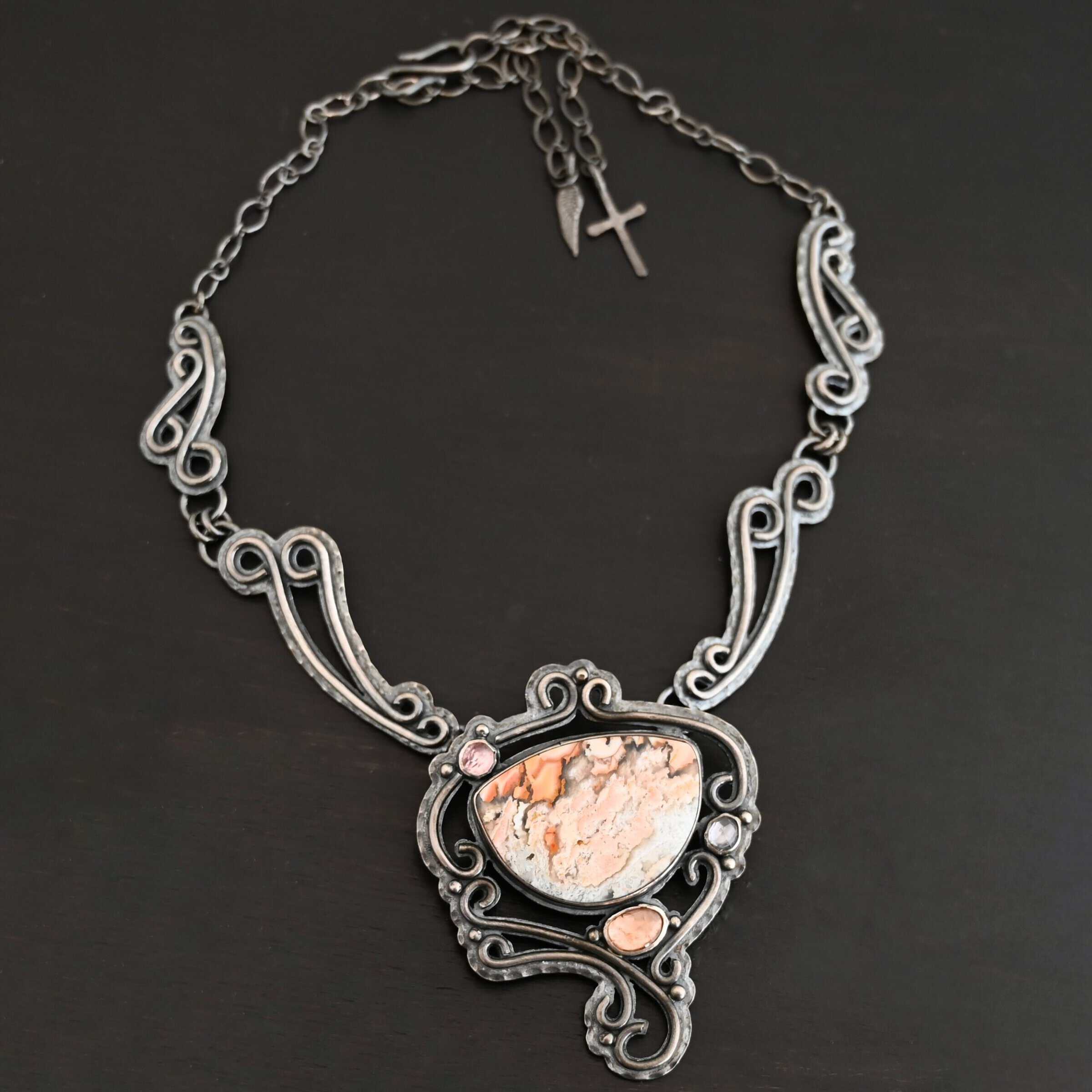 Meadow Wind Necklace New The "Meadow Wind" Necklace as shown close up. Necklace is made out of sterling silver, crazy lace agate, sapphires, and tourmaline. The agate is the main stone, and it is a slight oval shape. There are three other stones that are scattered around the necklace. The agate is surrounded by sterling silver swirls, and the collar of the necklace is made out of these swirls as well. Necklace is set against a dark background.
