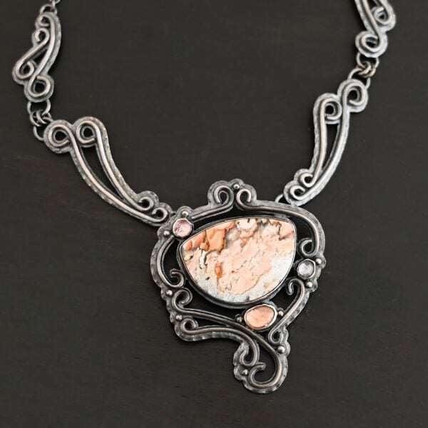 The "Meadow Wind" Necklace as shown close up. Necklace is made out of sterling silver, crazy lace agate, sapphires, and tourmaline. The agate is the main stone, and it is a slight oval shape. There are three other stones that are scattered around the necklace. The agate is surrounded by sterling silver swirls, and the collar of the necklace is made out of these swirls as well. Necklace is set against a dark background.