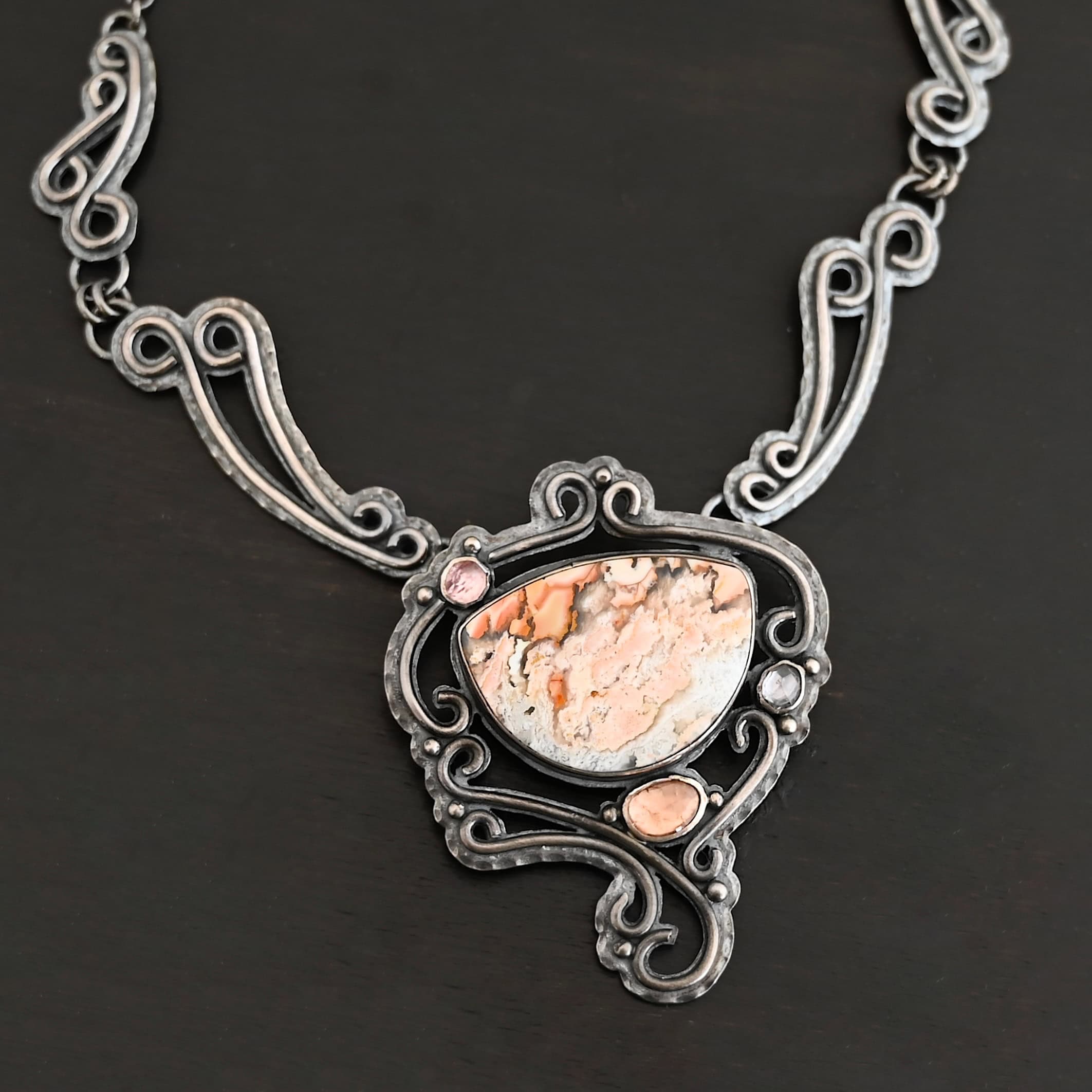 Meadow Wind Necklace New The "Meadow Wind" Necklace as shown close up. Necklace is made out of sterling silver, crazy lace agate, sapphires, and tourmaline. The agate is the main stone, and it is a slight oval shape. There are three other stones that are scattered around the necklace. The agate is surrounded by sterling silver swirls, and the collar of the necklace is made out of these swirls as well. Necklace is set against a dark background.