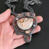 The "Meadow Wind" Necklace as shown close up. Necklace is made out of sterling silver, crazy lace agate, sapphires, and tourmaline. The agate is the main stone, and it is a slight oval shape. There are three other stones that are scattered around the necklace. The agate is surrounded by sterling silver swirls, and the collar of the necklace is made out of these swirls as well. Necklace is set against the palm of a hand to demonstrate size of the pendant and collar of the necklace.