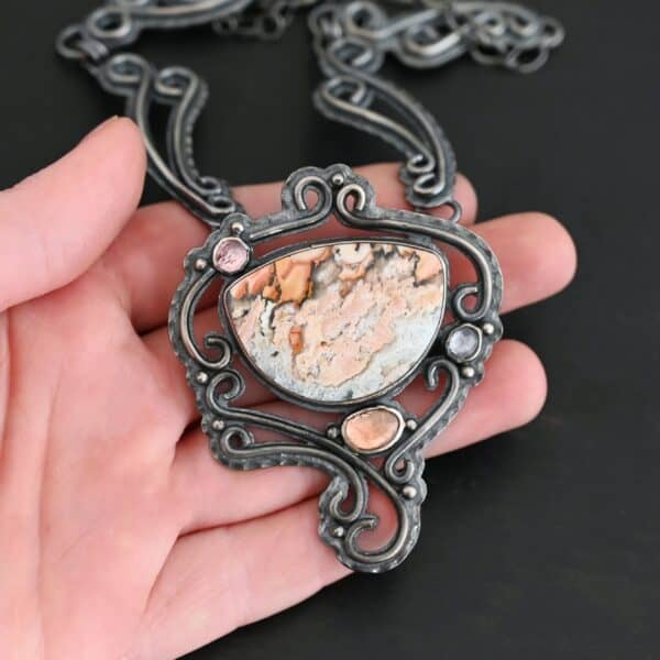 The "Meadow Wind" Necklace as shown close up. Necklace is made out of sterling silver, crazy lace agate, sapphires, and tourmaline. The agate is the main stone, and it is a slight oval shape. There are three other stones that are scattered around the necklace. The agate is surrounded by sterling silver swirls, and the collar of the necklace is made out of these swirls as well. Necklace is set against the palm of a hand to demonstrate size of the pendant and collar of the necklace.