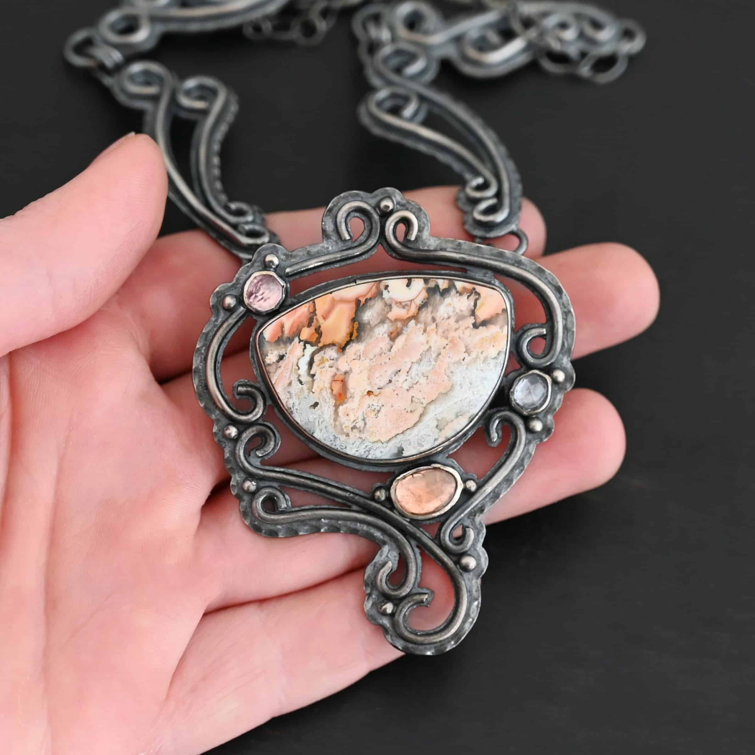 Meadow Wind Necklace New The "Meadow Wind" Necklace as shown close up. Necklace is made out of sterling silver, crazy lace agate, sapphires, and tourmaline. The agate is the main stone, and it is a slight oval shape. There are three other stones that are scattered around the necklace. The agate is surrounded by sterling silver swirls, and the collar of the necklace is made out of these swirls as well. Necklace is set against the palm of a hand to demonstrate size of the pendant and collar of the necklace.