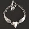 Close up of the "Courage" Bracelet. Bracelet is made out of solid sterling silver with a casted heart and two angel wings. Casted heart is solid sterling silver, and connected by an angel wing on each side. Angel wings are connected to a chain with a toggle clasp to connect the bracelet together. Bracelet is shiny and the heart is hammered to give it a textured look. Bracelet is set against a dark background.
