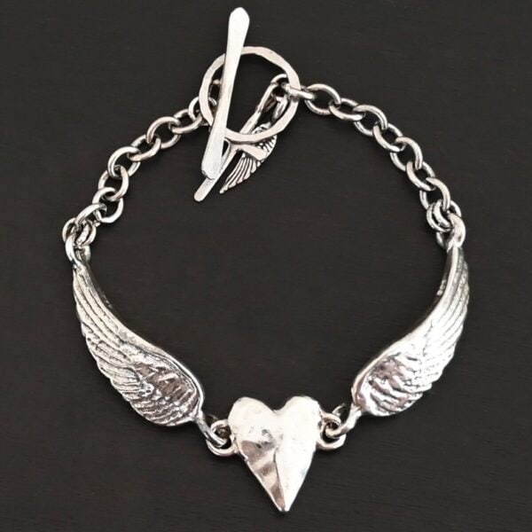 Close up of the "Courage" Bracelet. Bracelet is made out of solid sterling silver with a casted heart and two angel wings. Casted heart is solid sterling silver, and connected by an angel wing on each side. Angel wings are connected to a chain with a toggle clasp to connect the bracelet together. Bracelet is shiny and the heart is hammered to give it a textured look. Bracelet is set against a dark background.