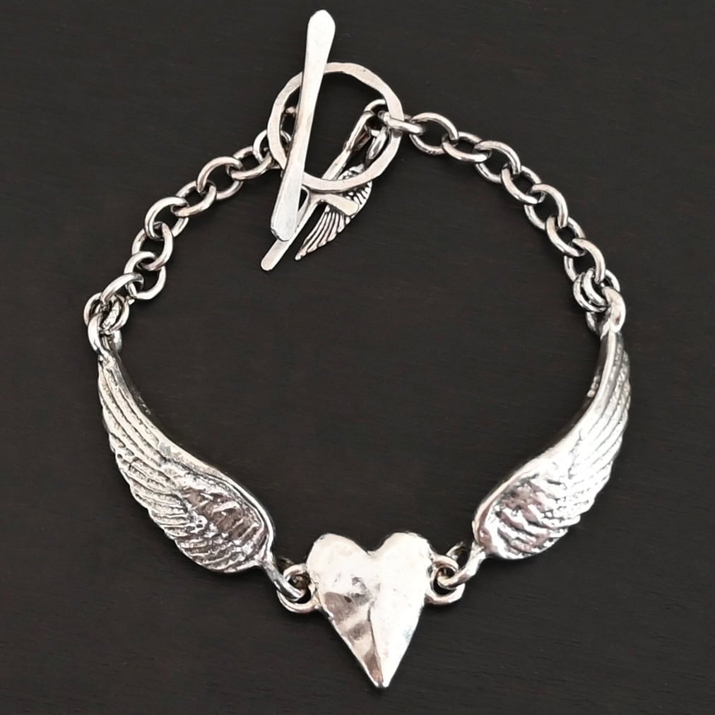 Courage Bracelet Close up of the "Courage" Bracelet. Bracelet is made out of solid sterling silver with a casted heart and two angel wings. Casted heart is solid sterling silver, and connected by an angel wing on each side. Angel wings are connected to a chain with a toggle clasp to connect the bracelet together. Bracelet is shiny and the heart is hammered to give it a textured look. Bracelet is set against a dark background.