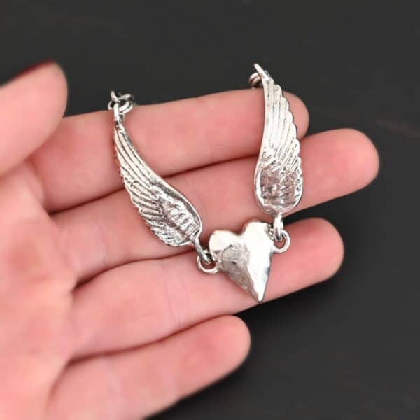 Close up of the "Courage" Bracelet. Bracelet is made out of solid sterling silver with a casted heart and two angel wings. Casted heart is solid sterling silver, and connected by an angel wing on each side. Angel wings are connected to a chain with a toggle clasp to connect the bracelet together. Bracelet is shiny and the heart is hammered to give it a textured look. Bracelet is set against a dark background.