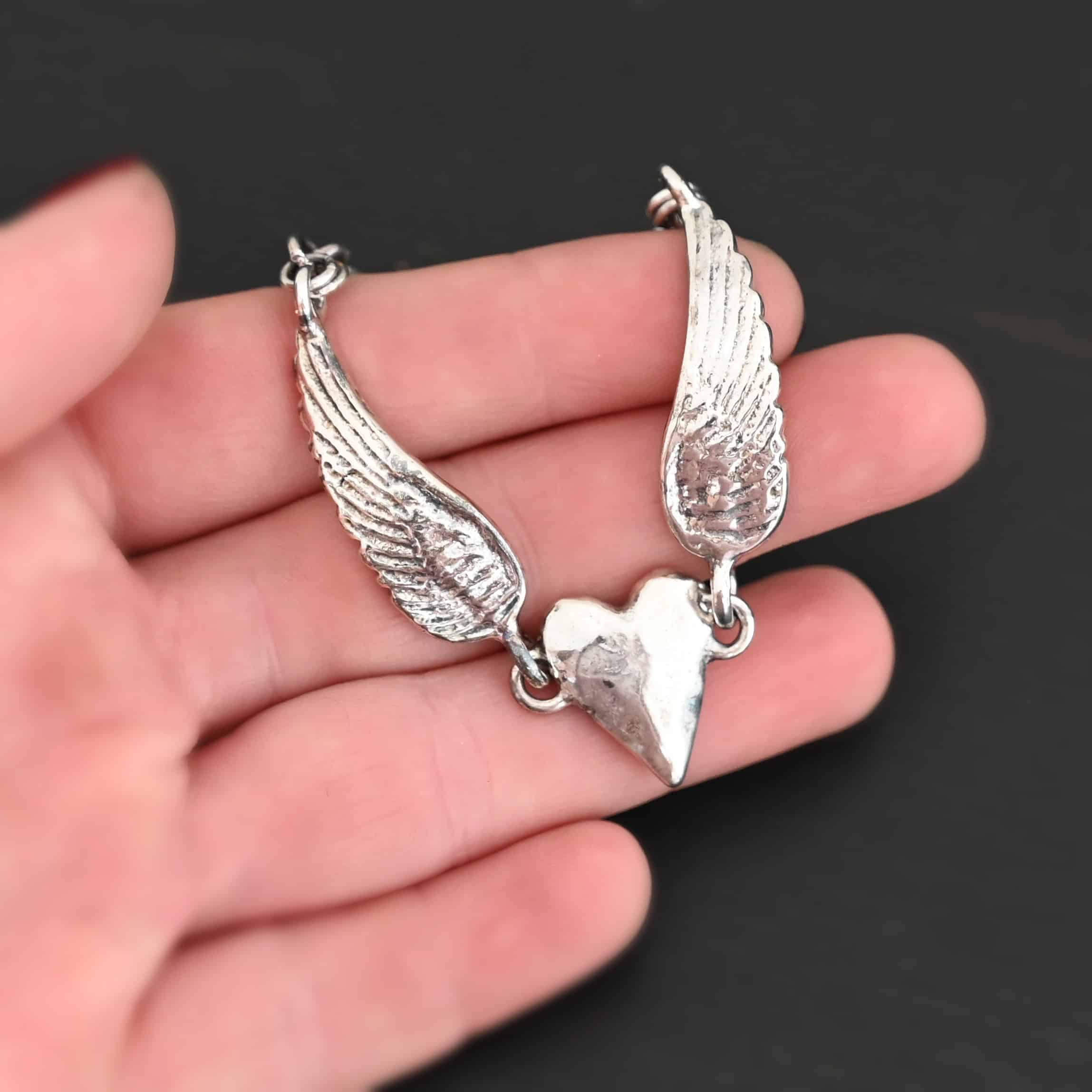 Courage Bracelet Close up of the "Courage" Bracelet. Bracelet is made out of solid sterling silver with a casted heart and two angel wings. Casted heart is solid sterling silver, and connected by an angel wing on each side. Angel wings are connected to a chain with a toggle clasp to connect the bracelet together. Bracelet is shiny and the heart is hammered to give it a textured look. Bracelet is set against a dark background.