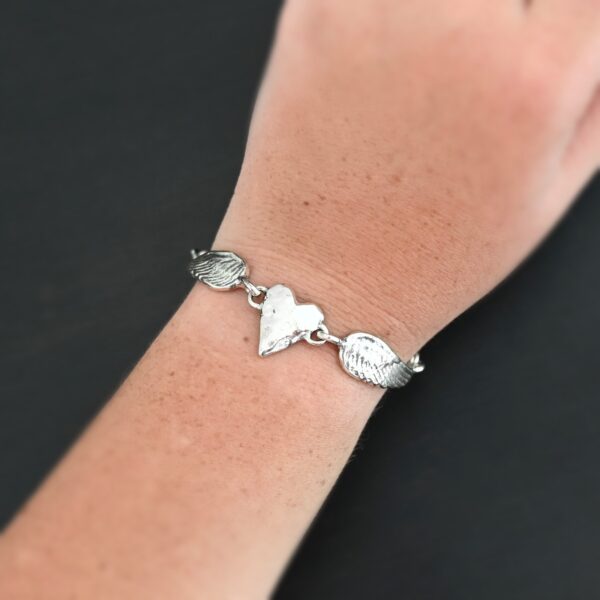Close up of the "Courage" Bracelet. Bracelet is made out of solid sterling silver with a casted heart and two angel wings. Casted heart is solid sterling silver, and connected by an angel wing on each side. Angel wings are connected to a chain with a toggle clasp to connect the bracelet together. Bracelet is shiny and the heart is hammered to give it a textured look. Bracelet is worn by a model to demonstrate size.