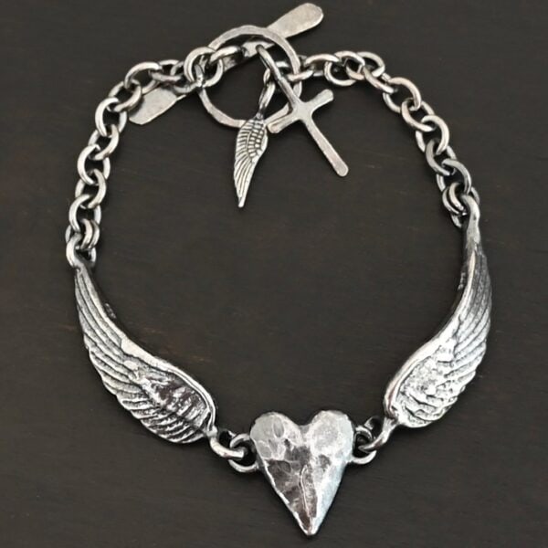 Close up of the "Courage" Bracelet. Bracelet is made out of solid sterling silver with a casted heart and two angel wings. Casted heart is solid sterling silver, and connected by an angel wing on each side. Angel wings are connected to a chain with a toggle clasp to connect the bracelet together. Bracelet is oxidized and the heart is hammered to give it a textured look. Bracelet is set against a dark background.
