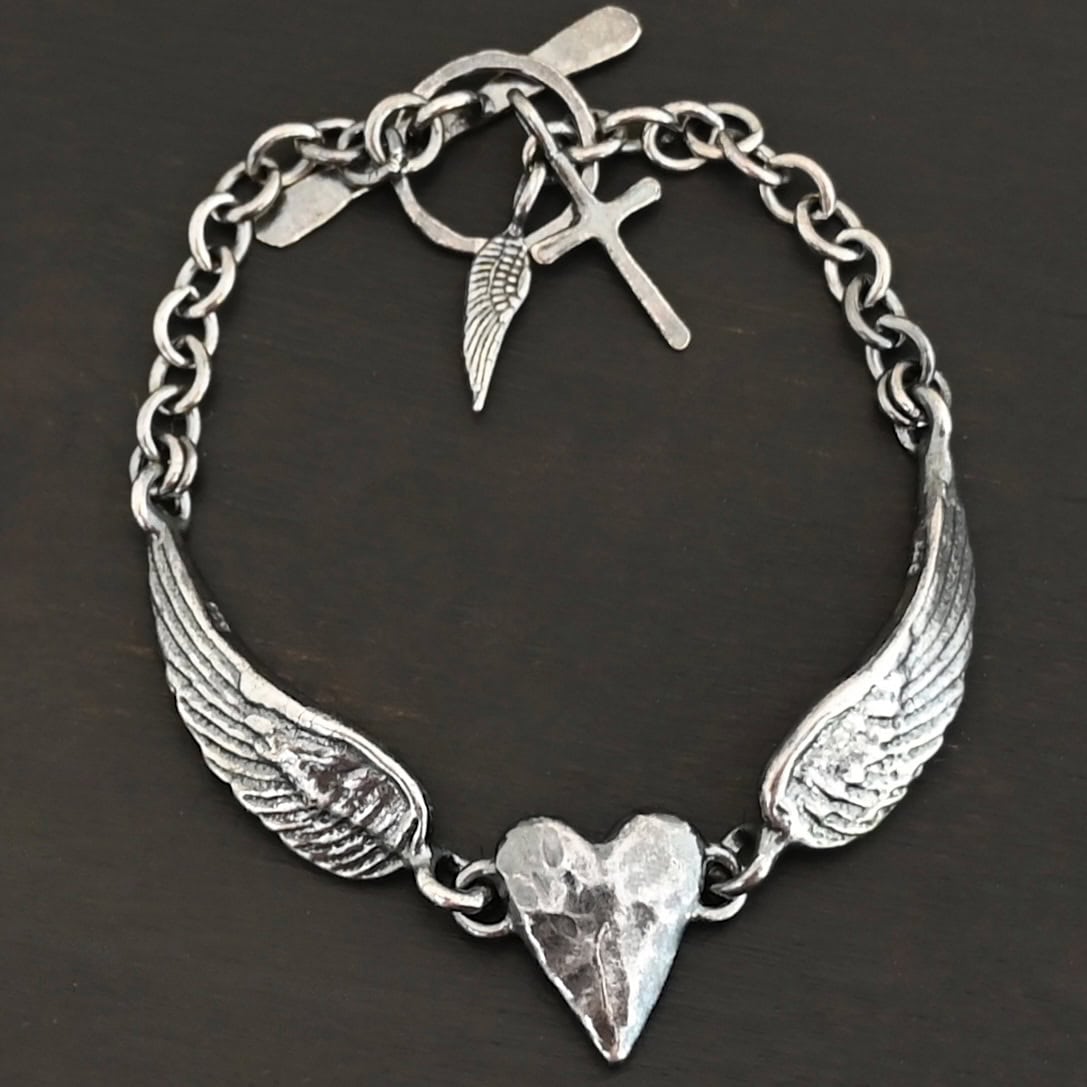 Courage Bracelet Close up of the "Courage" Bracelet. Bracelet is made out of solid sterling silver with a casted heart and two angel wings. Casted heart is solid sterling silver, and connected by an angel wing on each side. Angel wings are connected to a chain with a toggle clasp to connect the bracelet together. Bracelet is oxidized and the heart is hammered to give it a textured look. Bracelet is set against a dark background.