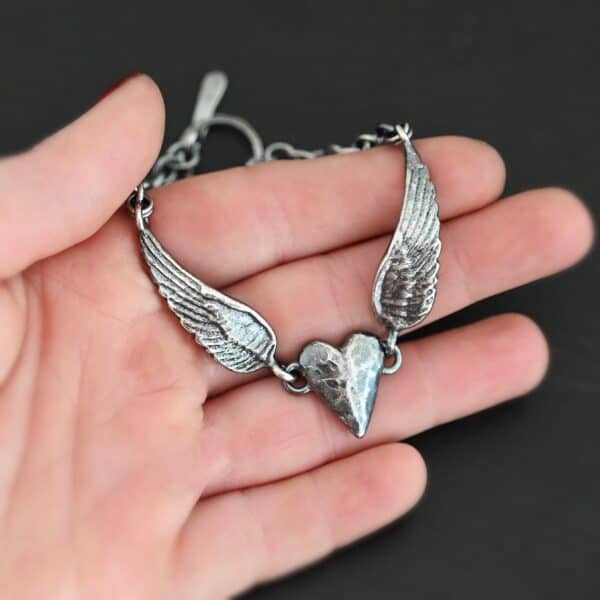Close up of the "Courage" Bracelet. Bracelet is made out of solid sterling silver with a casted heart and two angel wings. Casted heart is solid sterling silver, and connected by an angel wing on each side. Angel wings are connected to a chain with a toggle clasp to connect the bracelet together. Bracelet is oxidized and the heart is hammered to give it a textured look. Bracelet is set against a dark background.