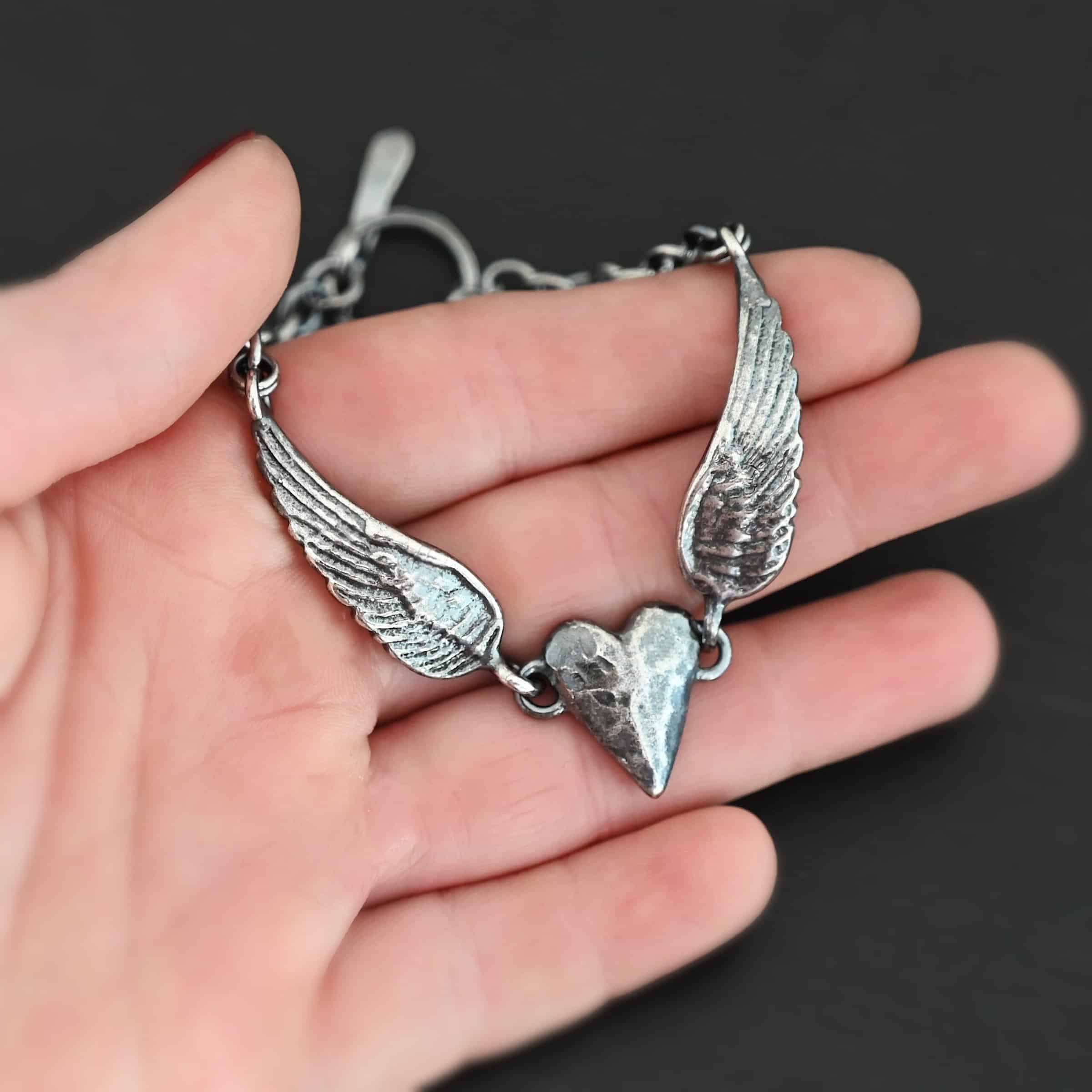 Courage Bracelet Close up of the "Courage" Bracelet. Bracelet is made out of solid sterling silver with a casted heart and two angel wings. Casted heart is solid sterling silver, and connected by an angel wing on each side. Angel wings are connected to a chain with a toggle clasp to connect the bracelet together. Bracelet is oxidized and the heart is hammered to give it a textured look. Bracelet is set against a dark background.