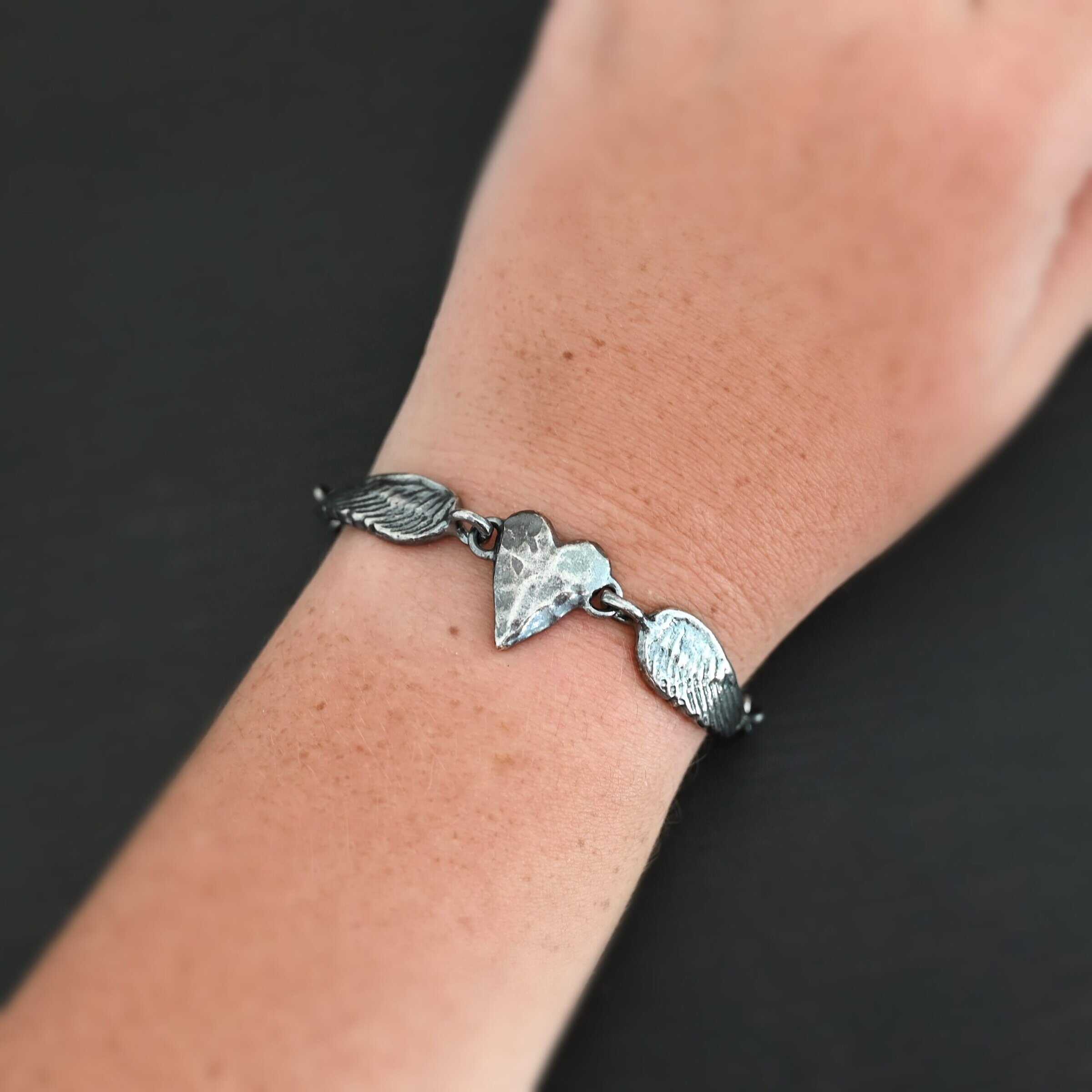 Courage Bracelet Close up of the "Courage" Bracelet. Bracelet is made out of solid sterling silver with a casted heart and two angel wings. Casted heart is solid sterling silver, and connected by an angel wing on each side. Angel wings are connected to a chain with a toggle clasp to connect the bracelet together. Bracelet is oxidized and the heart is hammered to give it a textured look. Bracelet is worn by a model to demonstrate size.