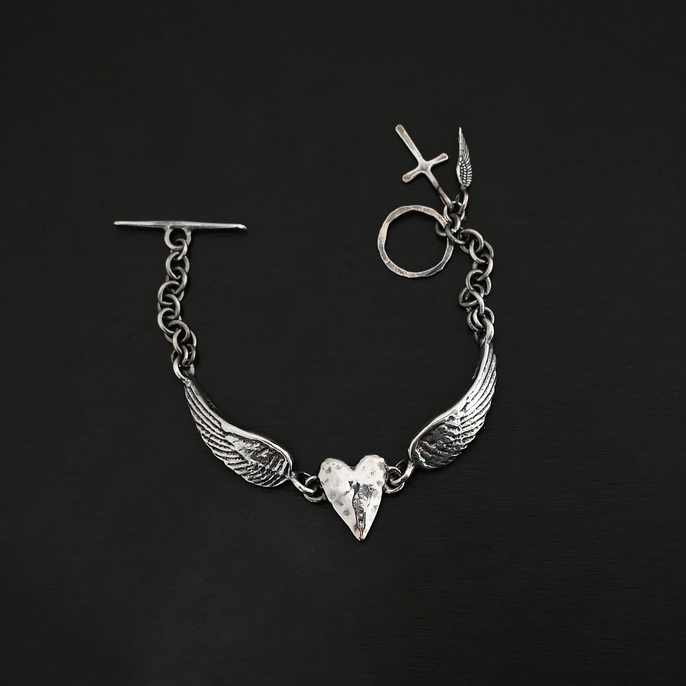 "Courage" Bracelet as shown in shiny. Bracelet is made out of solid sterling silver with a casted heart and two angel wings. Casted heart is solid sterling silver, and connected by an angel wing on each side. Angel wings are connected to a chain with a toggle clasp to connect the bracelet together. The heart is hammered to give it a textured look. Bracelet is set against a dark background.
