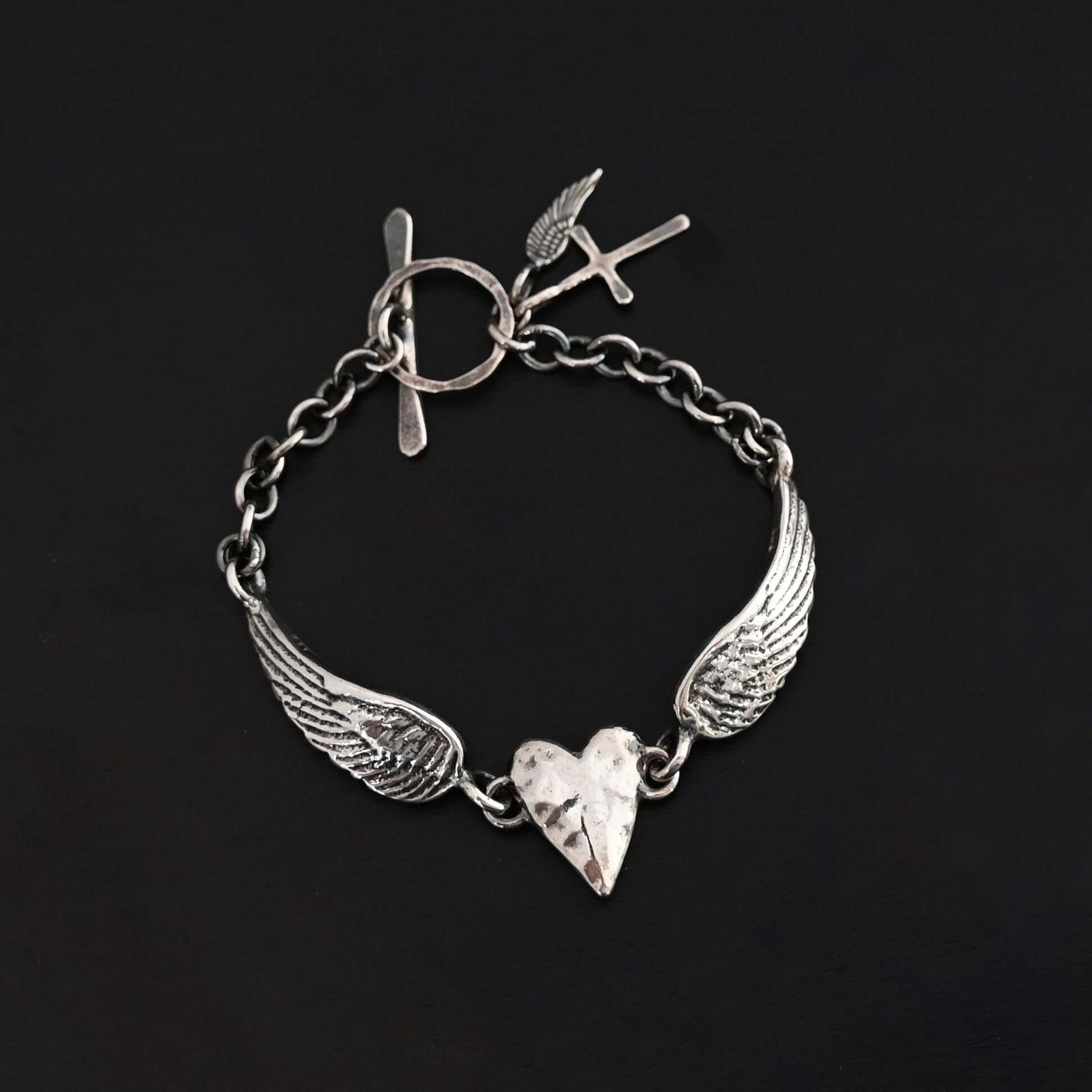 "Courage" Bracelet as shown in shiny. Bracelet is made out of solid sterling silver with a casted heart and two angel wings. Casted heart is solid sterling silver, and connected by an angel wing on each side. Angel wings are connected to a chain with a toggle clasp to connect the bracelet together. The heart is hammered to give it a textured look. Bracelet is shown against a dark background.
