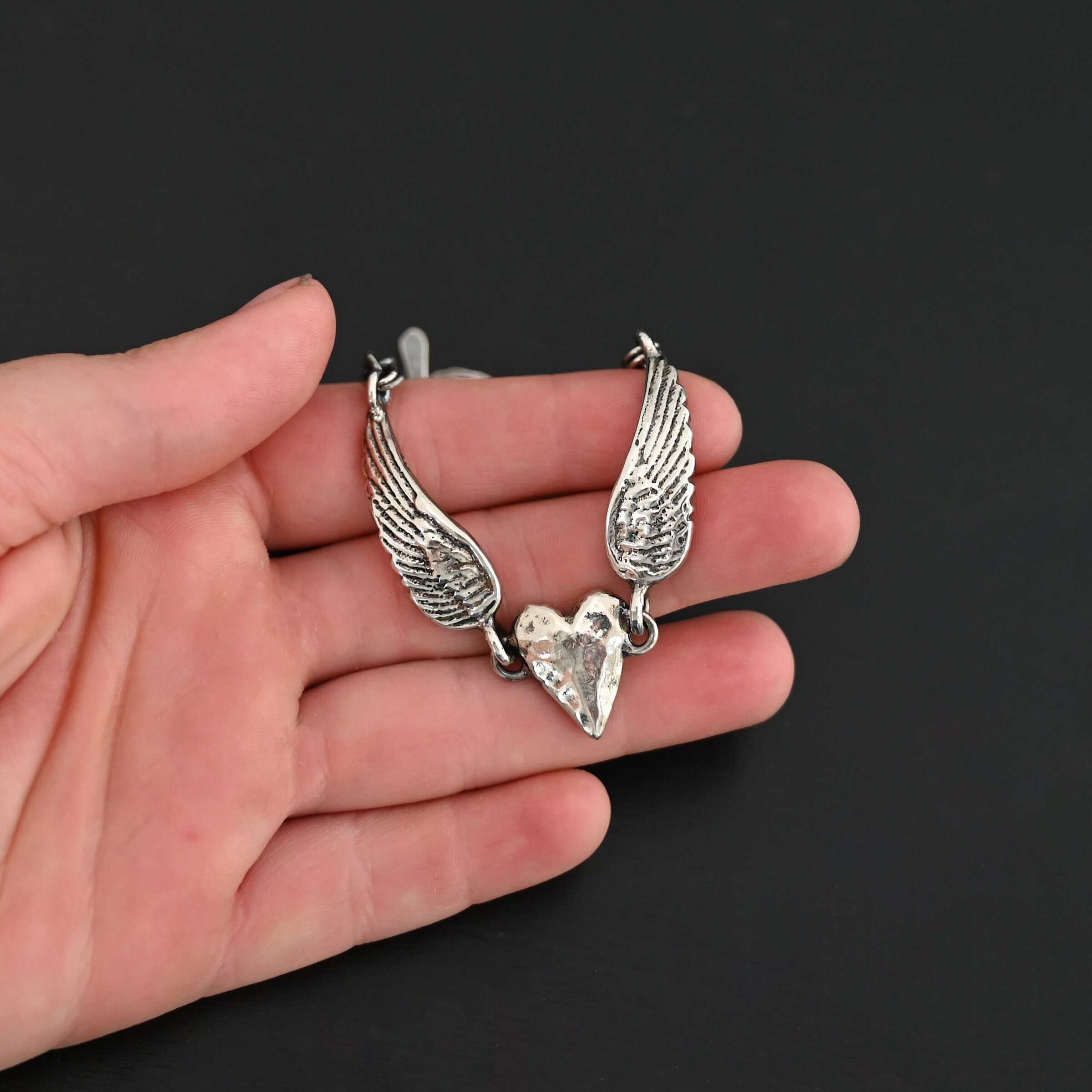 "Courage" Bracelet as shown in shiny. Bracelet is made out of solid sterling silver with a casted heart and two angel wings. Casted heart is solid sterling silver, and connected by an angel wing on each side. Angel wings are connected to a chain with a toggle clasp to connect the bracelet together. The heart is hammered to give it a textured look. Bracelet is shown against a models fingertips to demonstrate size.