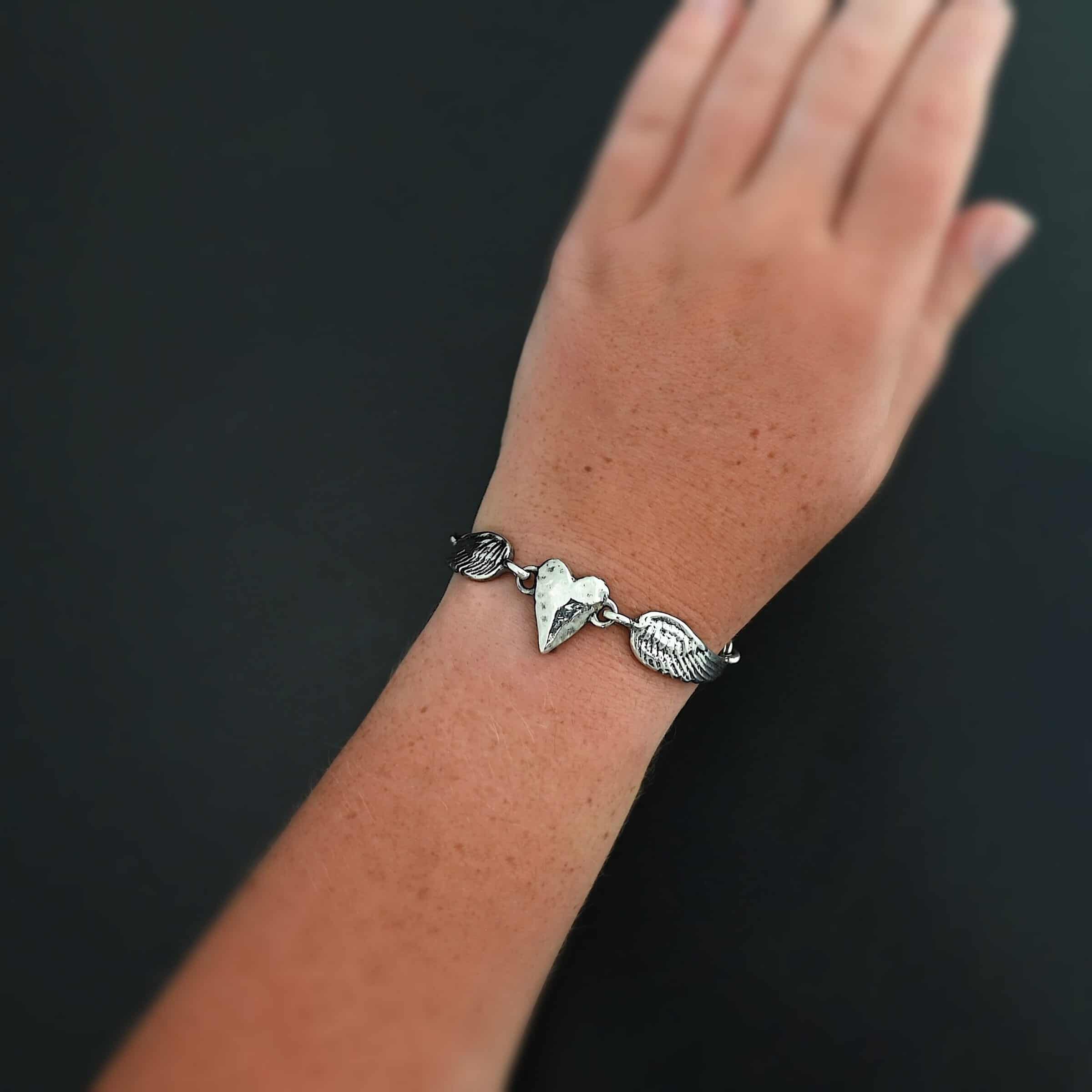 "Courage" Bracelet as shown in shiny. Bracelet is made out of solid sterling silver with a casted heart and two angel wings. Casted heart is solid sterling silver, and connected by an angel wing on each side. Angel wings are connected to a chain with a toggle clasp to connect the bracelet together. The heart is hammered to give it a textured look. Bracelet is worn by a model to demonstrate size.