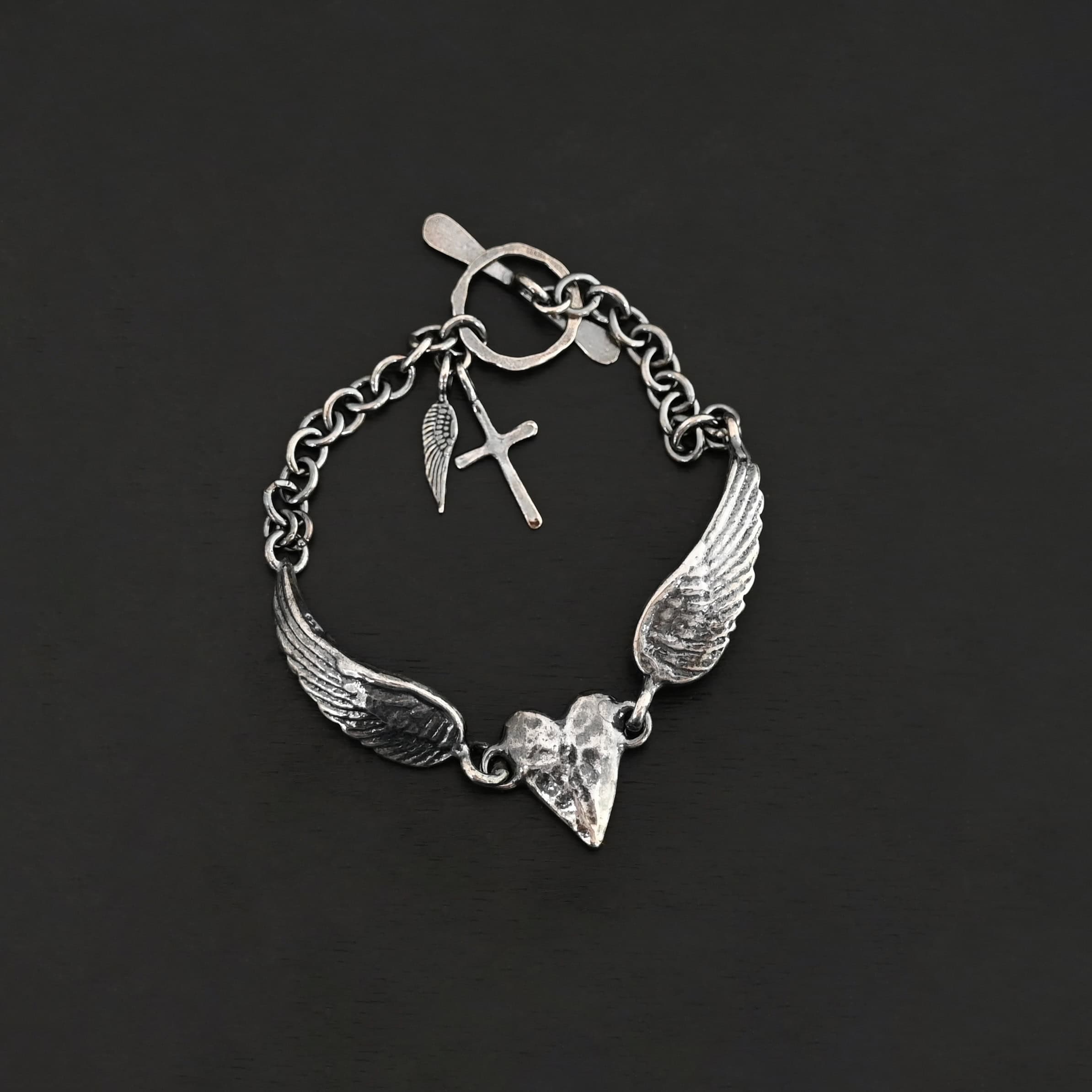 "Courage" Bracelet as shown in oxidized. Bracelet is made out of solid sterling silver with a casted heart and two angel wings. Casted heart is solid sterling silver, and connected by an angel wing on each side. Angel wings are connected to a chain with a toggle clasp to connect the bracelet together. The heart is hammered to give it a textured look. Bracelet is set against a dark background.