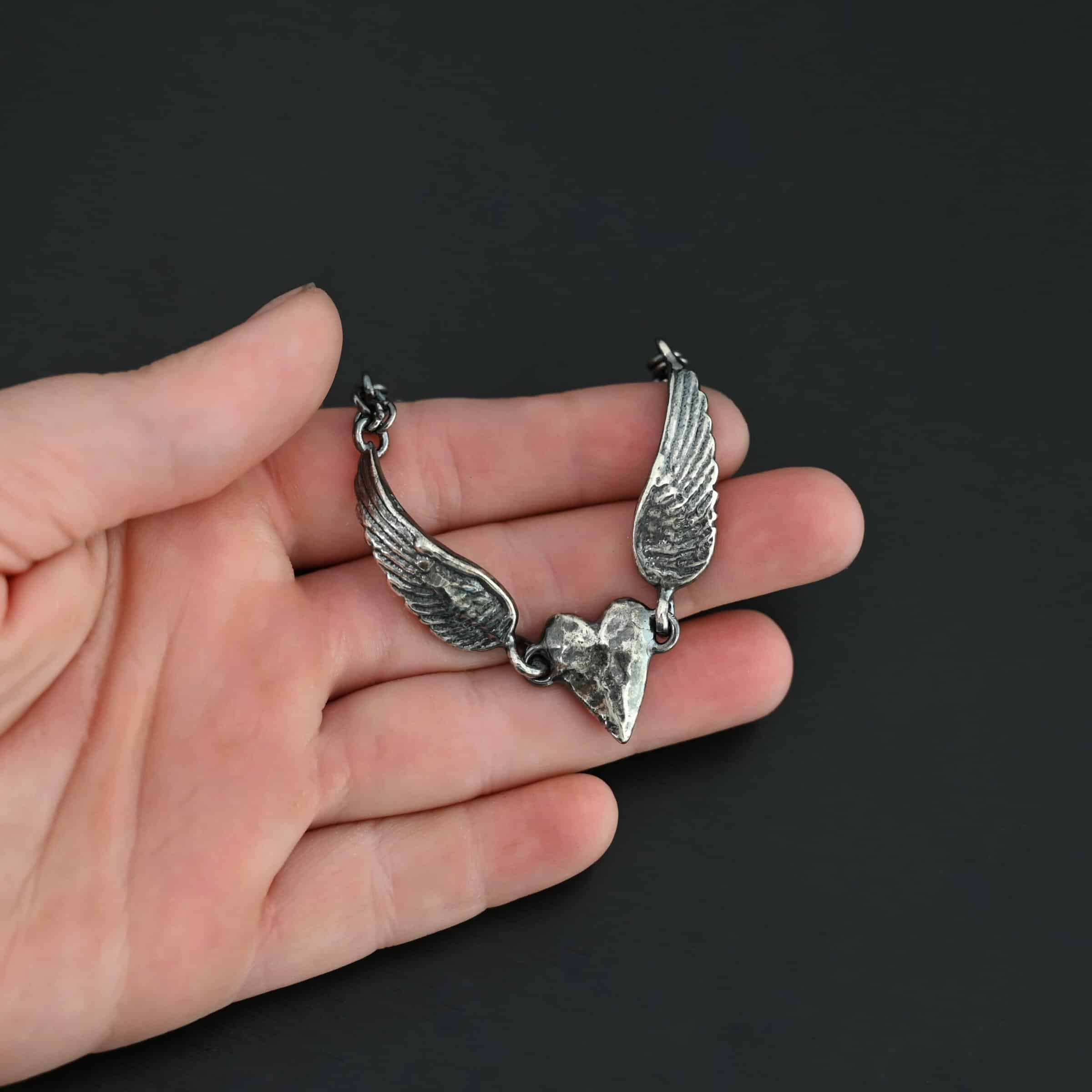 "Courage" Bracelet as shown in oxidized. Bracelet is made out of solid sterling silver with a casted heart and two angel wings. Casted heart is solid sterling silver, and connected by an angel wing on each side. Angel wings are connected to a chain with a toggle clasp to connect the bracelet together. The heart is hammered to give it a textured look. Bracelet is set against a models fingertips to demonstrate size.