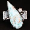 Photo of the "Just Add Water" Ring. There is a large, light blue, teardrop shaped larimar in the center of the large, square ring shank. The stone is set sideways, so the point of the teardrop is pointing towards the top left. There is a moissanite on the top right side of the square ring shank. There are four balls of silver, two on the right side and two on the left side of the ring shank. The larimar has six silver prongs holding it in, four on the bottom and two on the top.