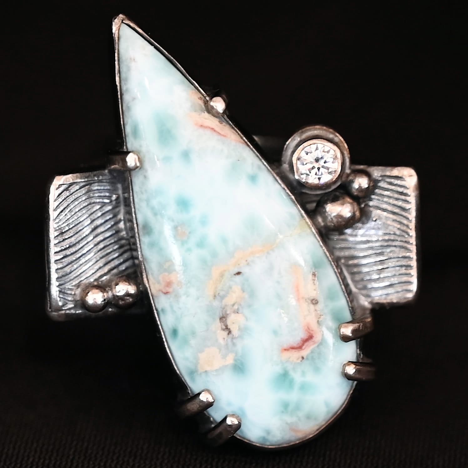 Just Add Water Ring Photo of the "Just Add Water" Ring. There is a large, light blue, teardrop shaped larimar in the center of the large, square ring shank. The stone is set sideways, so the point of the teardrop is pointing towards the top left. There is a moissanite on the top right side of the square ring shank. There are four balls of silver, two on the right side and two on the left side of the ring shank. The larimar has six silver prongs holding it in, four on the bottom and two on the top.