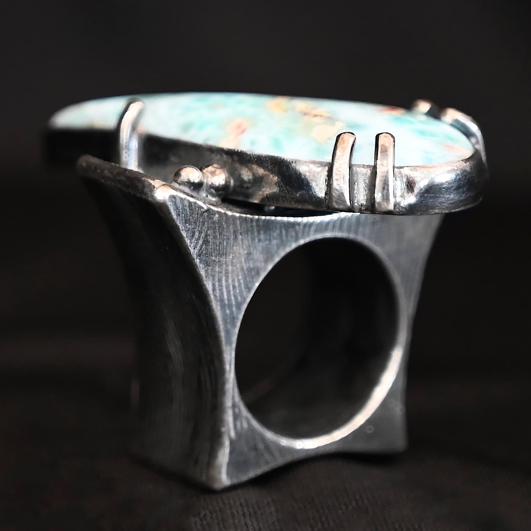 Just Add Water Ring Photo of the "Just Add Water" Ring. There is a large, light blue, teardrop shaped larimar in the center of the large, square ring shank. The stone is set sideways, so the point of the teardrop is pointing towards the top left. There is a moissanite on the top right side of the square ring shank. There are four balls of silver, two on the right side and two on the left side of the ring shank. The larimar has six silver prongs holding it in, four on the bottom and two on the top.