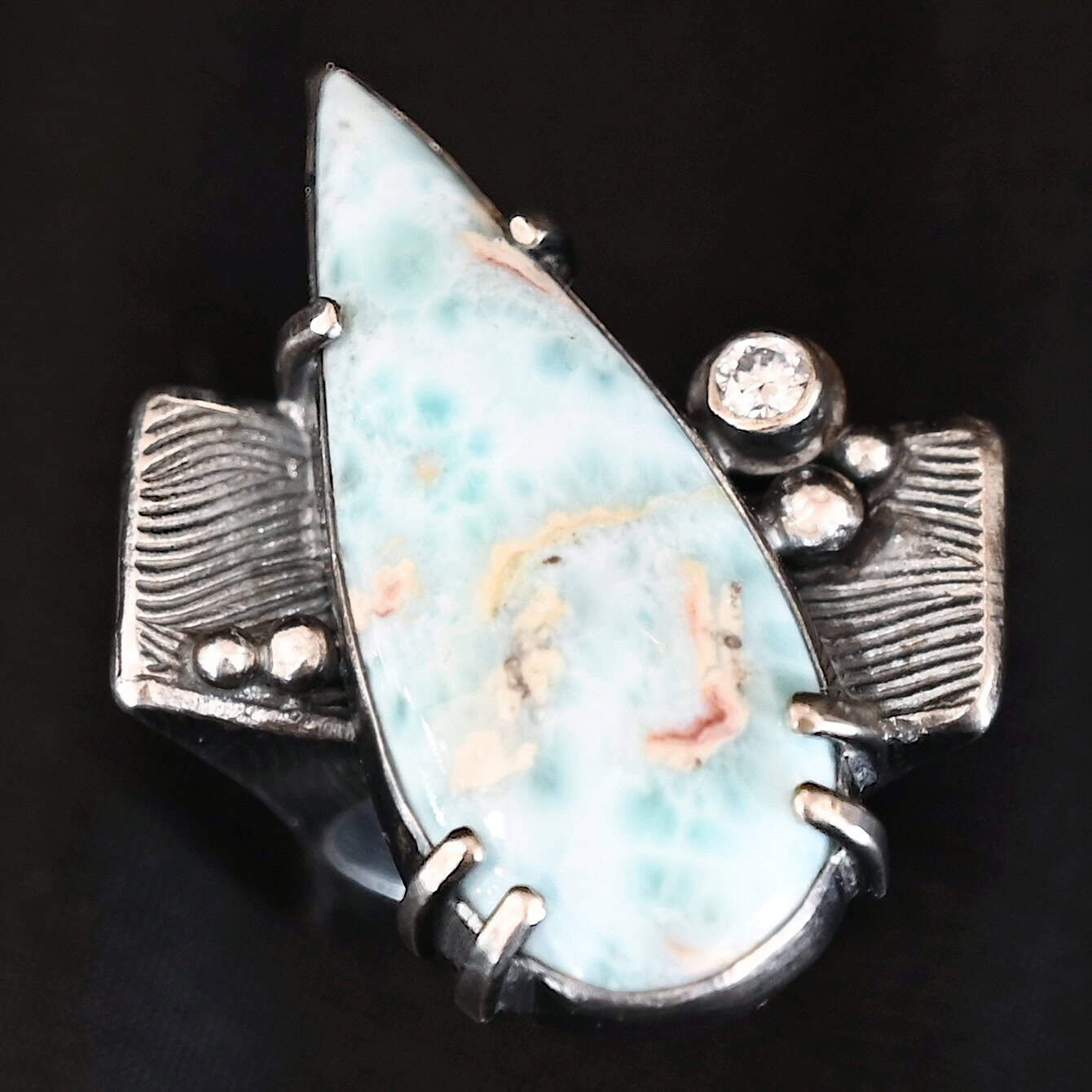Just Add Water Ring Photo of the "Just Add Water" Ring. There is a large, light blue, teardrop shaped larimar in the center of the large, square ring shank. The stone is set sideways, so the point of the teardrop is pointing towards the top left. There is a moissanite on the top right side of the square ring shank. There are four balls of silver, two on the right side and two on the left side of the ring shank. The larimar has six silver prongs holding it in, four on the bottom and two on the top.