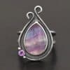 Close up of the "Elliana" Ring. Made out of sterling silver and fluorite and amethyst. The fluorite is the main stone in a teardrop shape. There is an amethyst in the bottom left corner of the ring. The silver comes up to the top to create two loops that are connected, one is taller than the other. The ring is resting against a dark background.