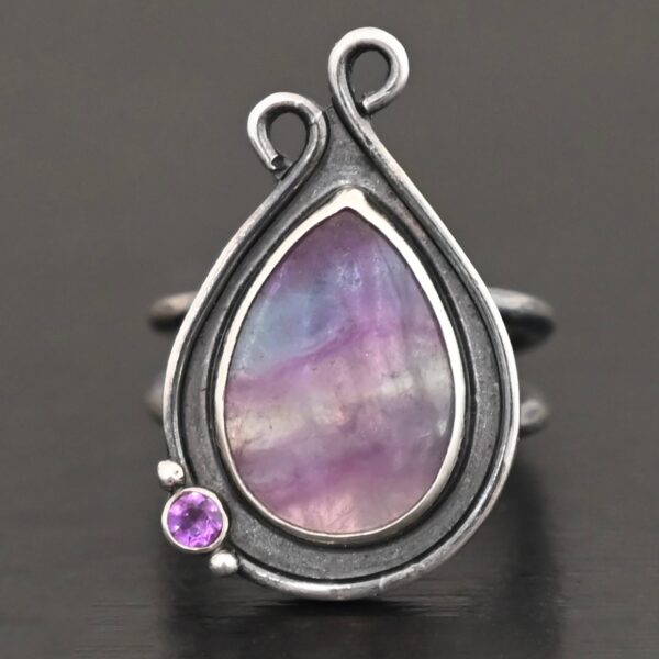 Close up of the "Elliana" Ring. Made out of sterling silver and fluorite and amethyst. The fluorite is the main stone in a teardrop shape. There is an amethyst in the bottom left corner of the ring. The silver comes up to the top to create two loops that are connected, one is taller than the other. The ring is resting against a dark background.