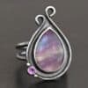 Close up of the "Elliana" Ring. Made out of sterling silver and fluorite and amethyst. The fluorite is the main stone in a teardrop shape. There is an amethyst in the bottom left corner of the ring. The silver comes up to the top to create two loops that are connected, one is taller than the other. The ring is resting against a dark background.