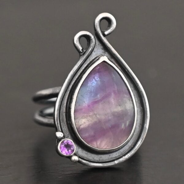 Close up of the "Elliana" Ring. Made out of sterling silver and fluorite and amethyst. The fluorite is the main stone in a teardrop shape. There is an amethyst in the bottom left corner of the ring. The silver comes up to the top to create two loops that are connected, one is taller than the other. The ring is resting against a dark background.