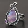 Close up of the "Elliana" Ring. Made out of sterling silver and fluorite and amethyst. The fluorite is the main stone in a teardrop shape. There is an amethyst in the bottom left corner of the ring. The silver comes up to the top to create two loops that are connected, one is taller than the other. The ring is resting against a dark background.